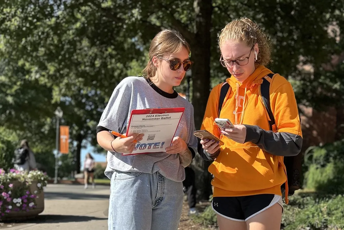 Two students stand outside and one holds a 2024 Election November Ballot clipboard